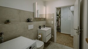 Un baño moderno con bañera, lavamanos e inodoro. Hay un espejo sobre el lavamanos y una puerta que da a otra habitación.