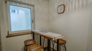 Una habitación pequeña con ventana, una mesa de madera cubierta con tela blanca, dos taburetes de madera y un reloj de pared.