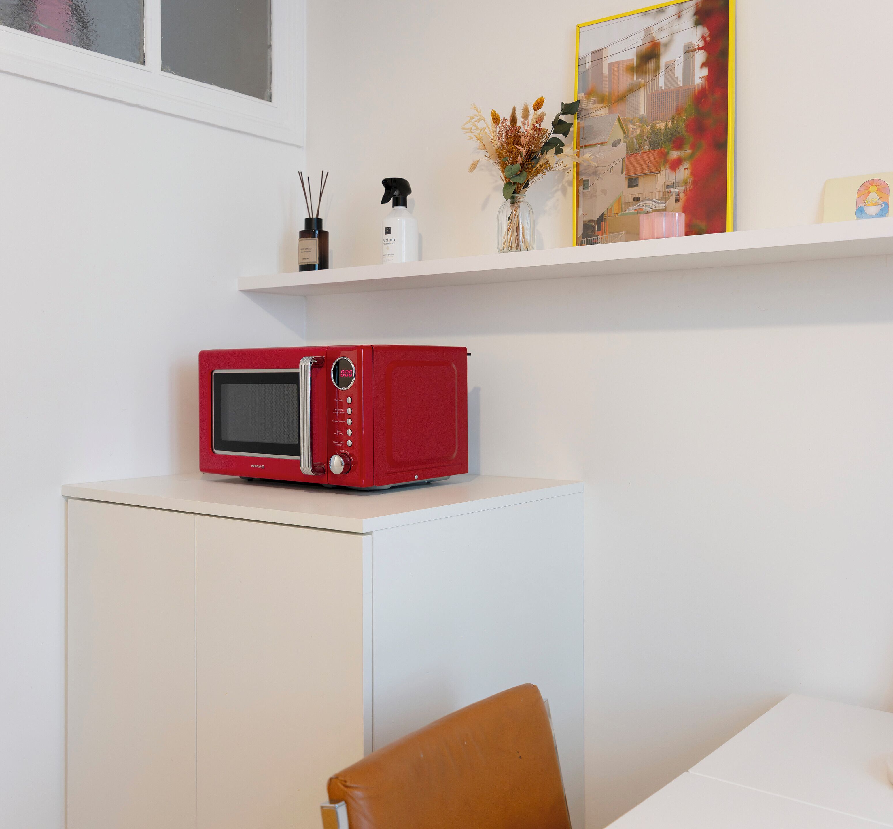 Un four à micro-ondes rouge posé sur un meuble blanc, une chaise marron et une étagère ornée d’objets décoratifs.