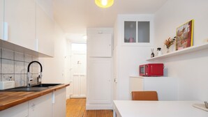 A compact kitchen with white cabinetry, a wooden countertop, a red microwave, and a white dining table with a brown chair. - Studio Secundo (Metz)