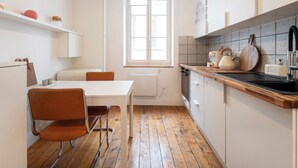 A compact kitchen with a wooden floor, white cabinetry, a dining table with two chairs, and a window providing natural light. - Studio Secundo (Metz)