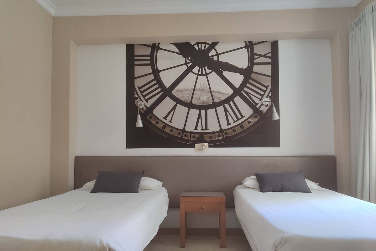 Une chambre d’hôtel avec deux lits simples, une petite table de chevet en bois et une grande horloge murale.