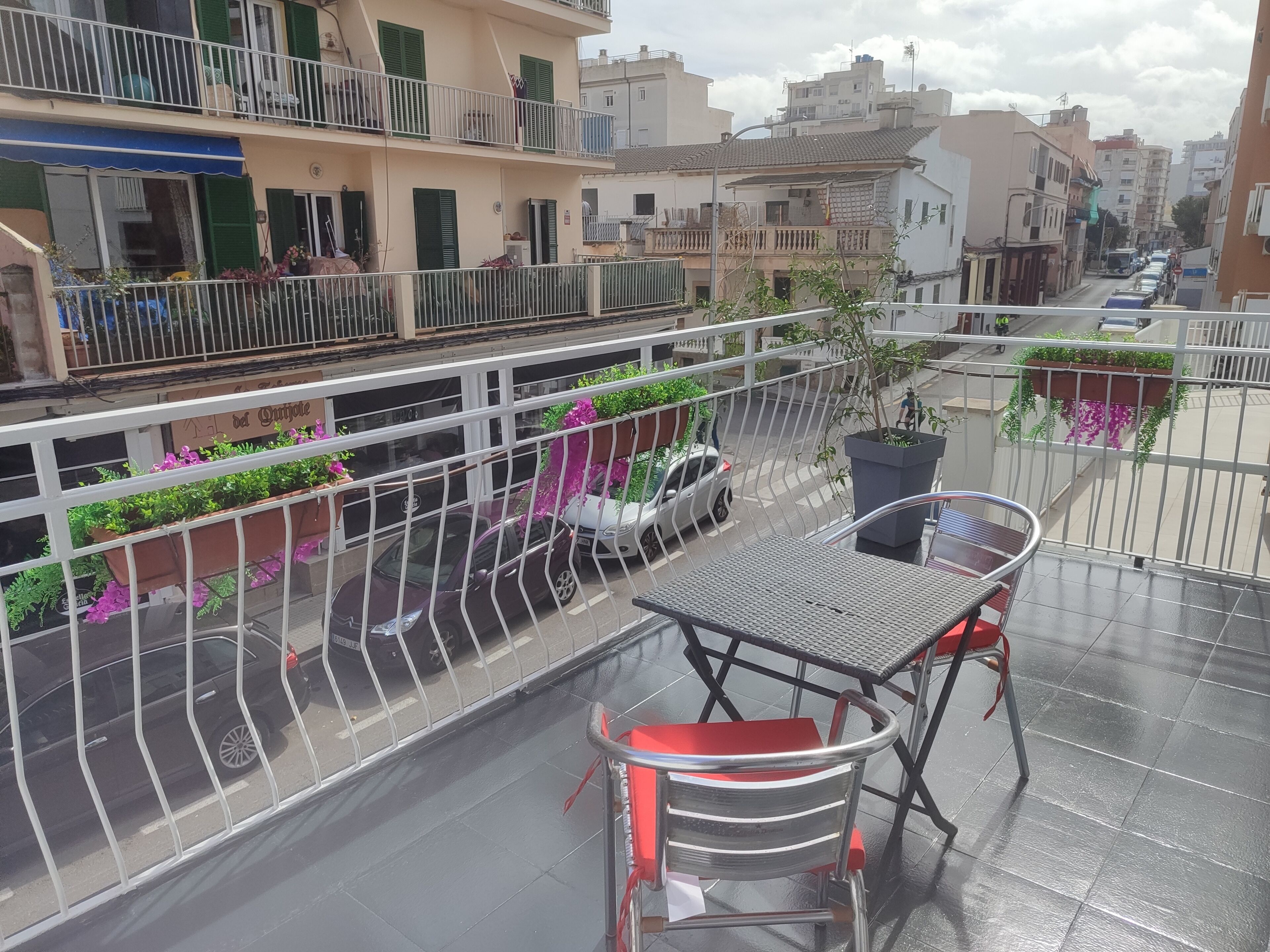A balcony with a metal railing, a table, and chairs. There are potted plants and a view of the street below.