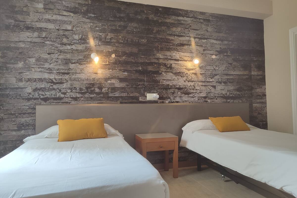 Une chambre d’hôtel avec deux lits, chacun doté de draps blancs et d’un seul oreiller jaune. La chambre est agrémentée d’un mur à texture évoquant le bois et de deux lampes fixées au mur.