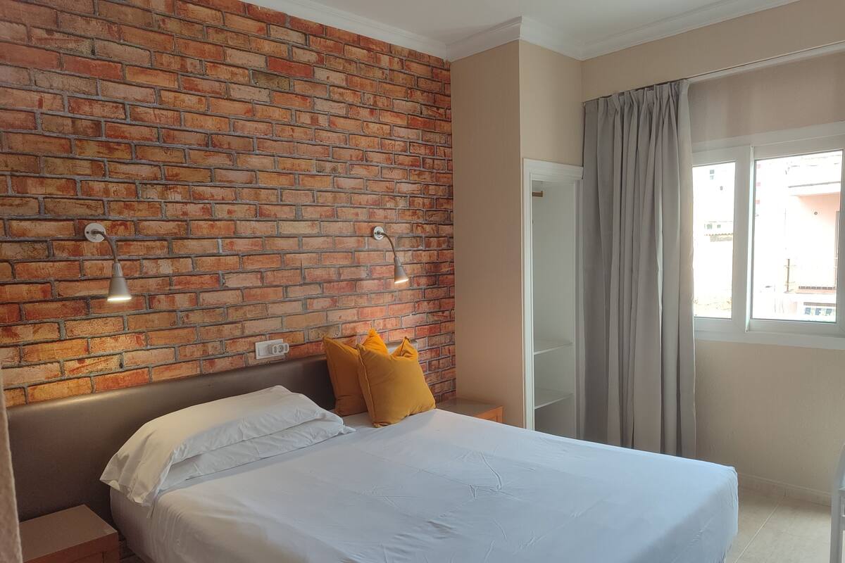Une chambre d’hôtel avec un mur en briques, un lit avec une literie blanche et des oreillers jaunes, une table de chevet, une fenêtre avec des rideaux et une lampe fixée au mur.