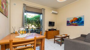 A living room with a sofa, a TV, a dining table with chairs, and a painting on the wall. - Apartment 'Musa Giardini Naxos' with Terrace, Garden and Air Conditioning (Giardini Naxos)