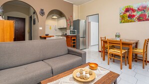A modern living room with a sofa, dining table, chairs, and a kitchen area with a refrigerator, oven, and microwave. - Apartment 'Musa Giardini Naxos' with Terrace, Garden and Air Conditioning (Giardini Naxos)