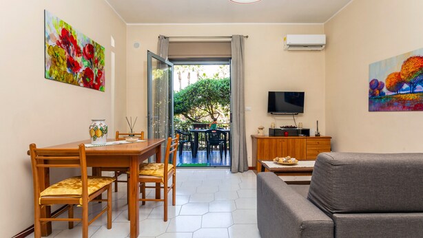 A dining area with a table and chairs, a sofa, a TV, and artwork on the walls. - Apartment 'Musa Giardini Naxos' with Terrace, Garden and Air Conditioning (Giardini Naxos)