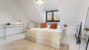A modern bedroom with a sloped ceiling, a large bed with white and orange bedding, a glass bedside table, a white chair, and a window with a view of trees. - Stuttgart Lux Living |Stylish Living in the Center (Stuttgart)