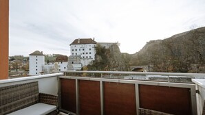 Ein Balkon mit Korbmöbeln, mit Blick auf eine Stadt mit einem markanten, schlossähnlichen Gebäude und einer Brücke.
