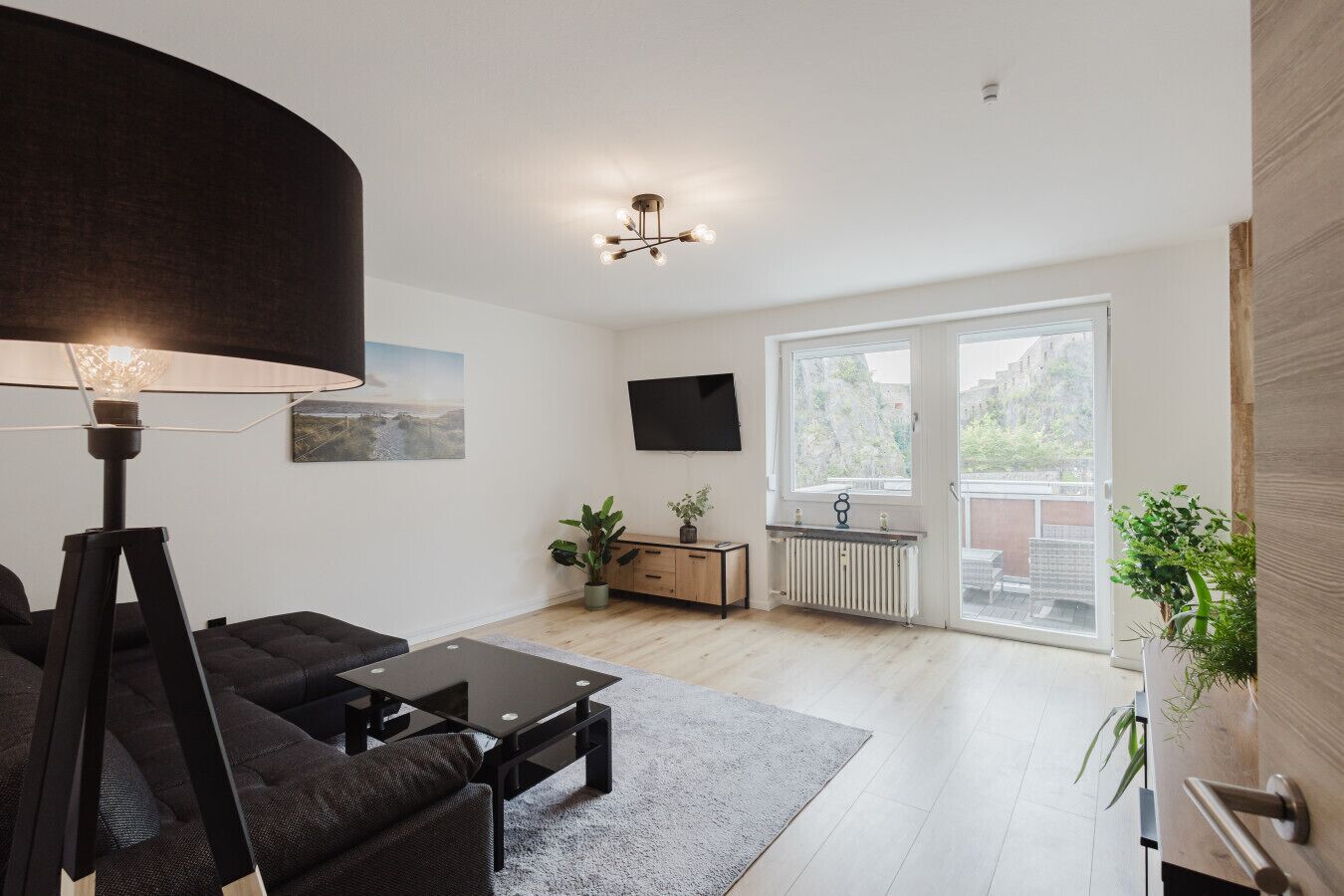 A modern living room with a sofa, coffee table, and lamp. There is a TV mounted on the wall, a radiator, and a window with a view of the outside.