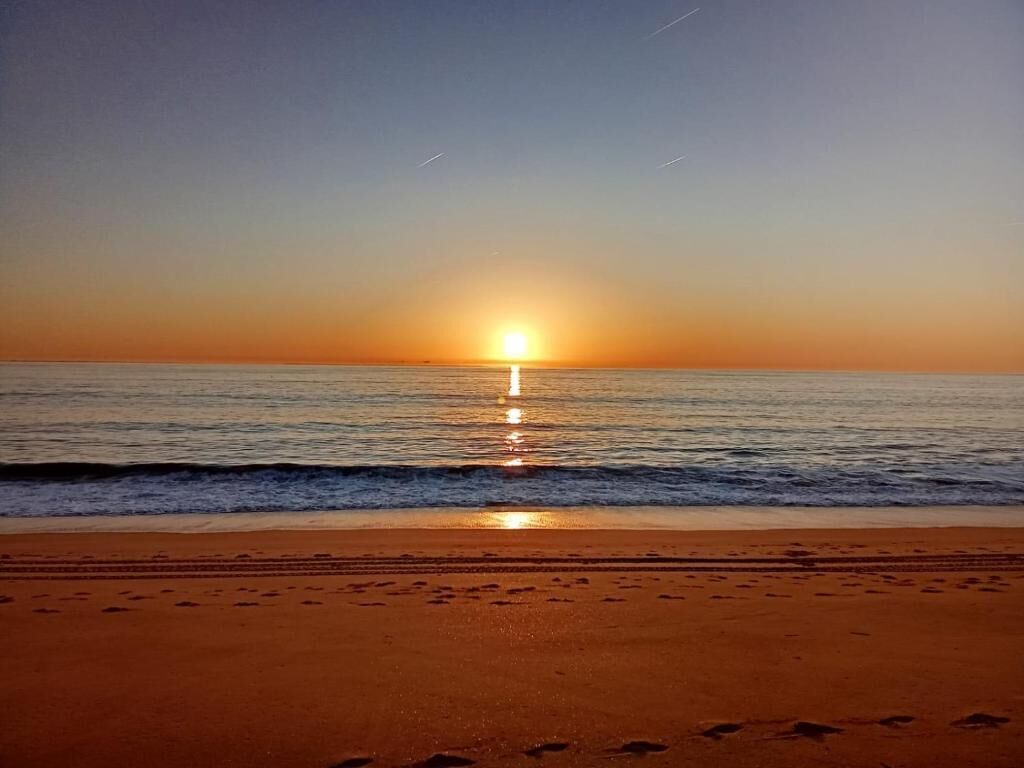 A beach with footprints leading to the water, a sunset over the horizon, and a calm sea.