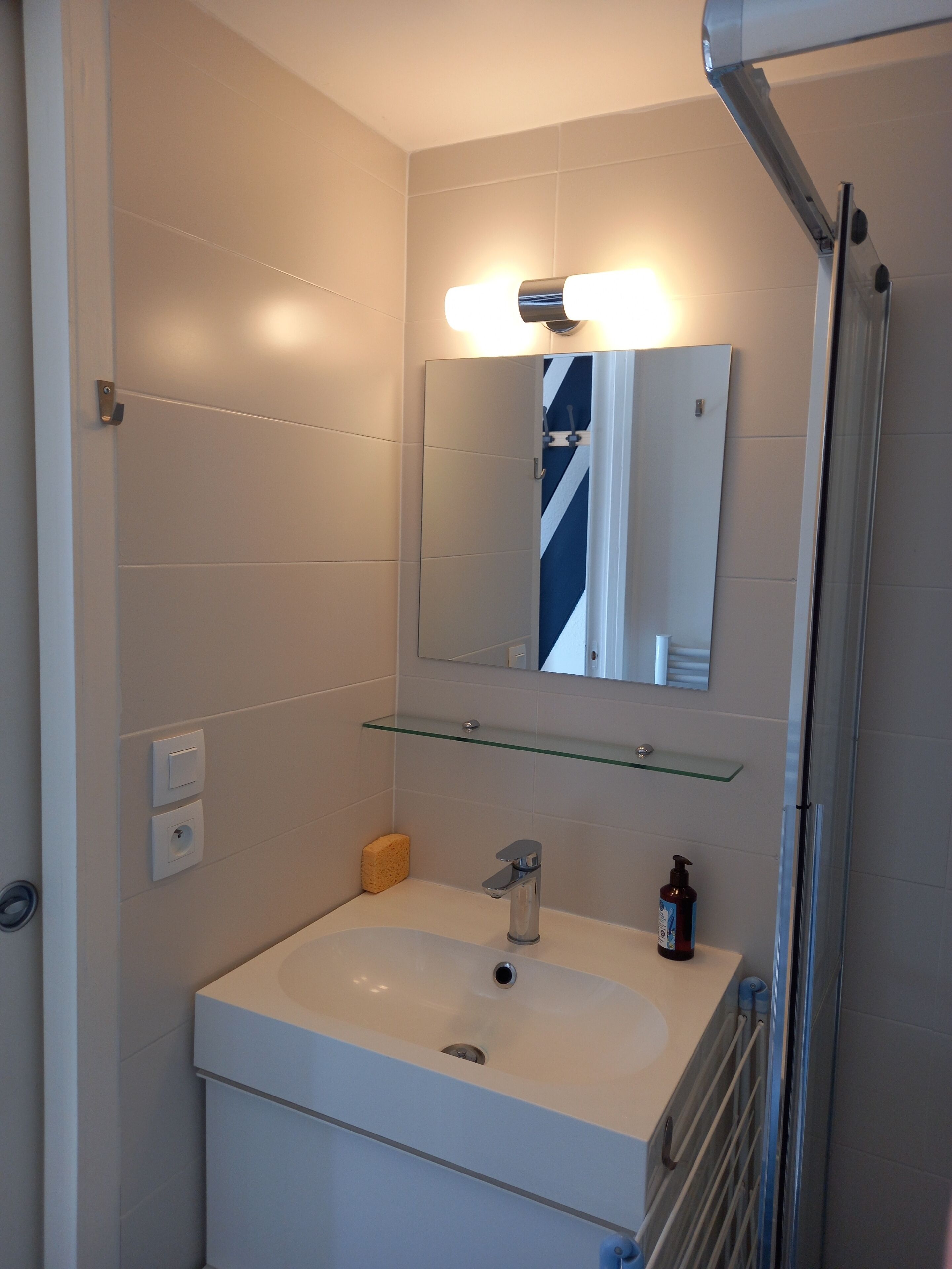 A modern bathroom with a white sink, a glass shelf above the sink, a mirror, and a shower area.
