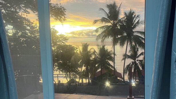 A view from a room with a glass door to a patio area. The patio has a tiled floor and a white chair. Palm trees and a building with a red roof are visible in the background. - Perla Pacifico (Puerto Armuelles)
