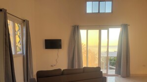 A spacious living room with a large sofa, a flat-screen TV mounted on the wall, and a sliding glass door leading to a balcony with a view of the sea. - Perla Pacifico (Puerto Armuelles)