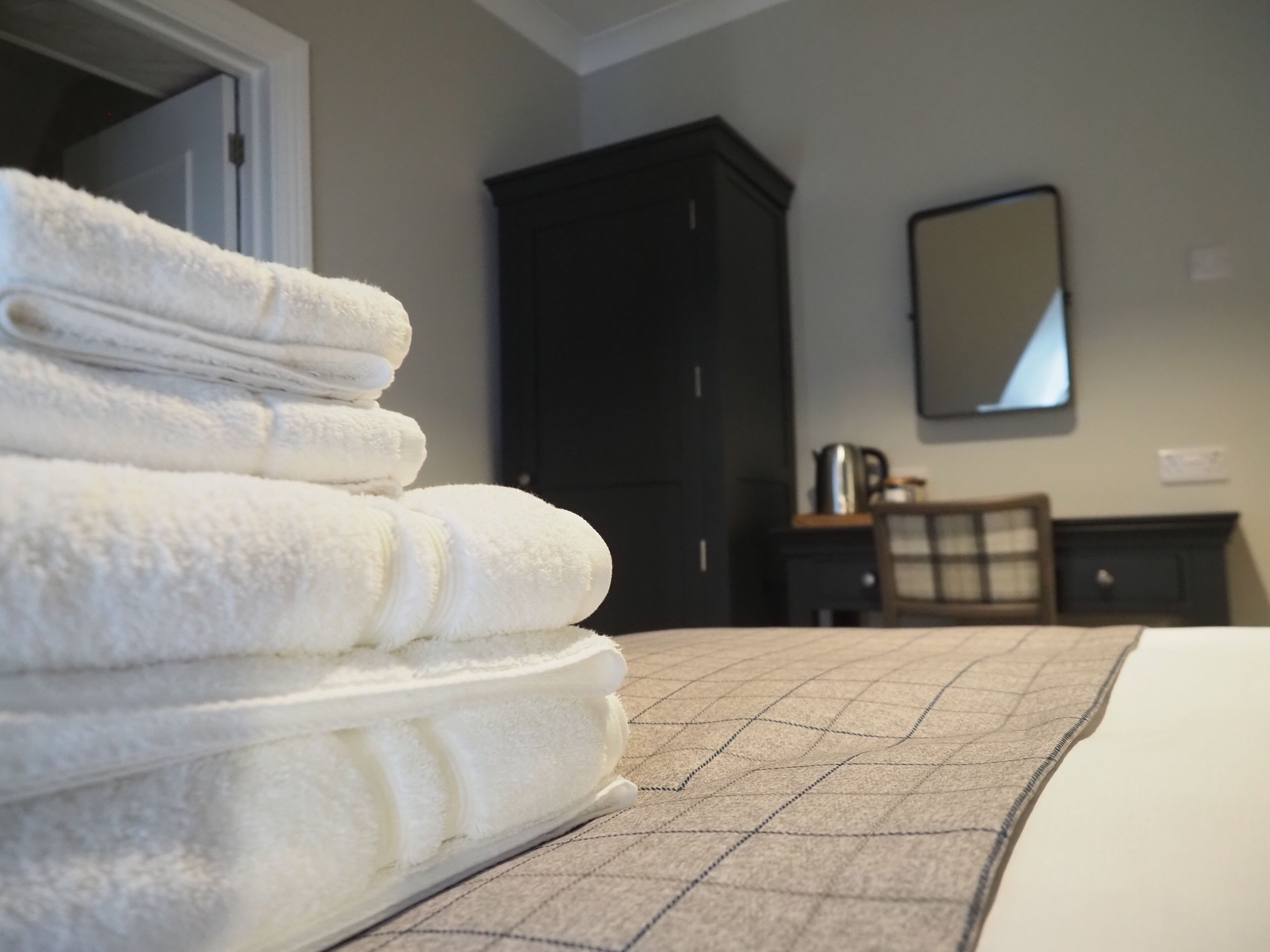 A neatly made bed with a checkered bedspread, a stack of white towels, a dark wooden wardrobe, and a mirror on the wall.