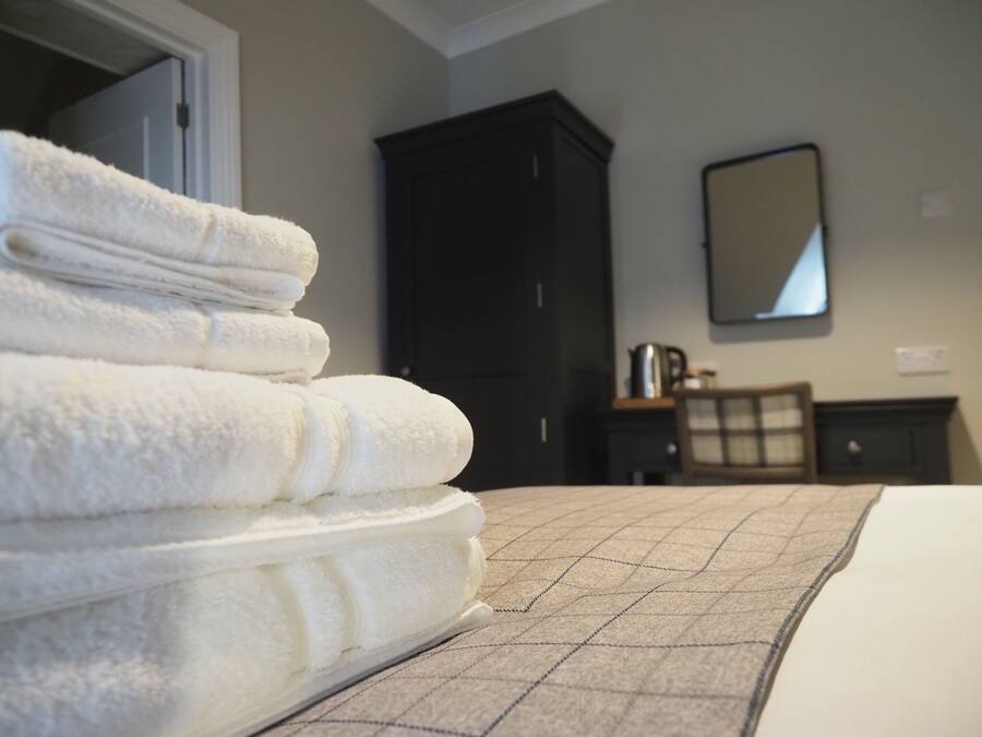 A neatly made bed with a checkered bedspread, a stack of white towels, a dark wooden wardrobe, and a mirror on the wall.