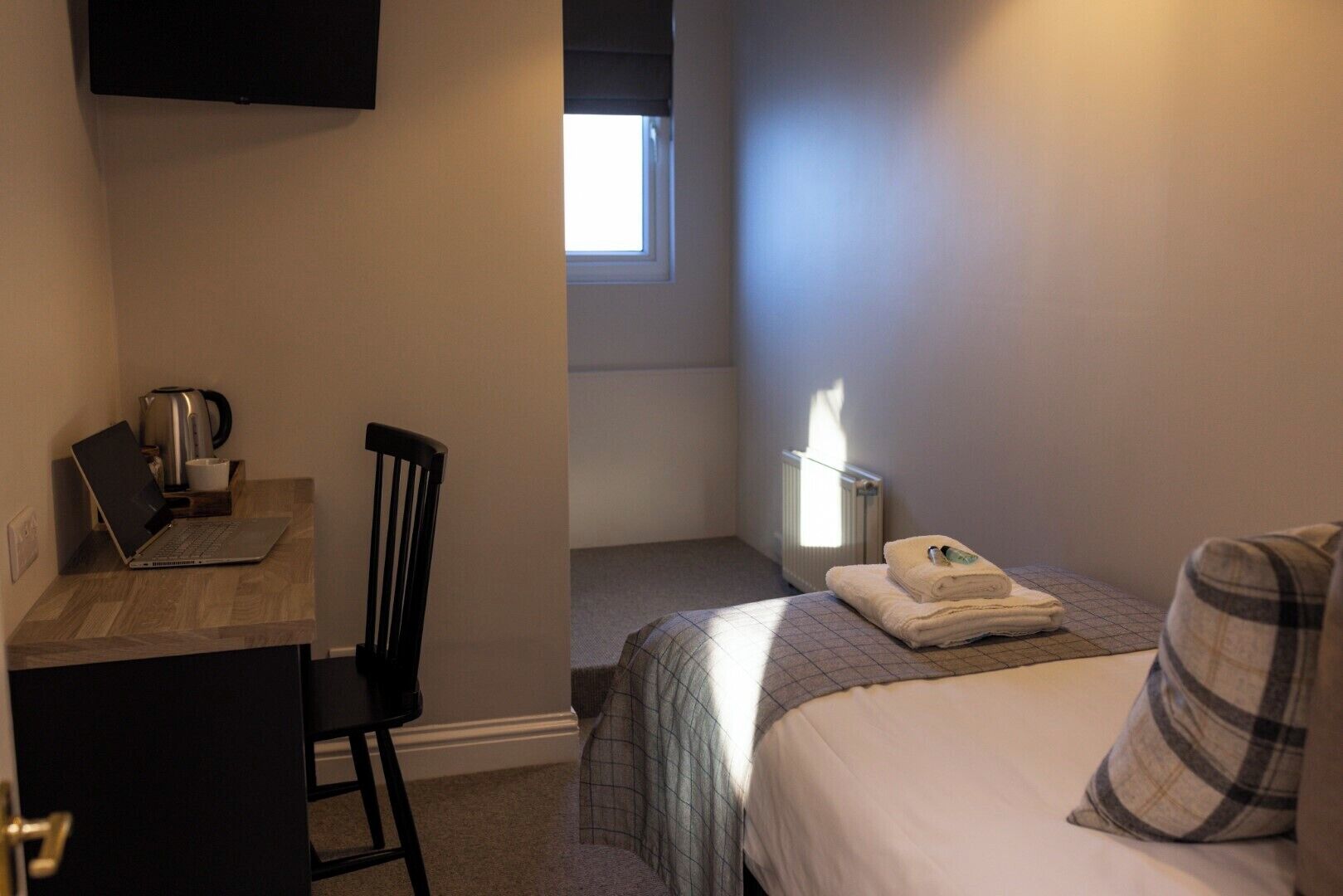 Une chambre d’hôtel de taille réduite, comprenant un lit, un bureau avec un ordinateur portable, une chaise, un radiateur et une fenêtre.