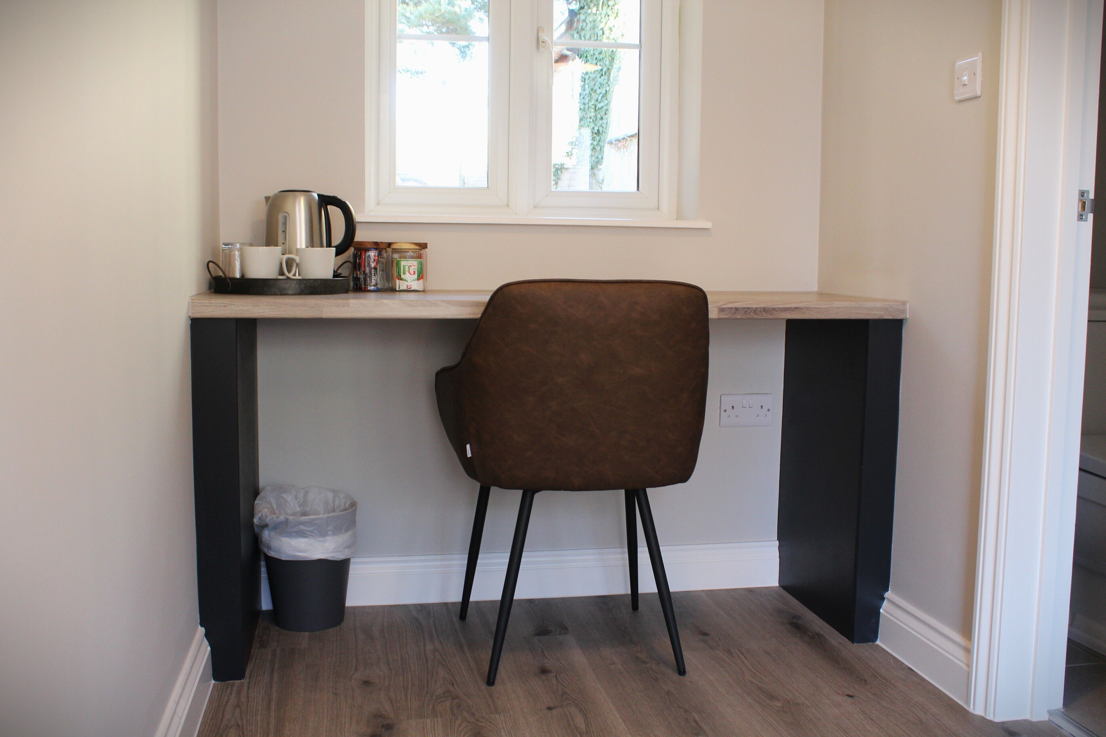 A small kitchen area with a wooden countertop, a brown chair, a trash bin, and a window with a view of trees.