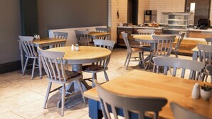 A modern restaurant with round tables and grey chairs, a kitchen area in the background, and a tiled floor. - The Pavilion Bisley (Woking)