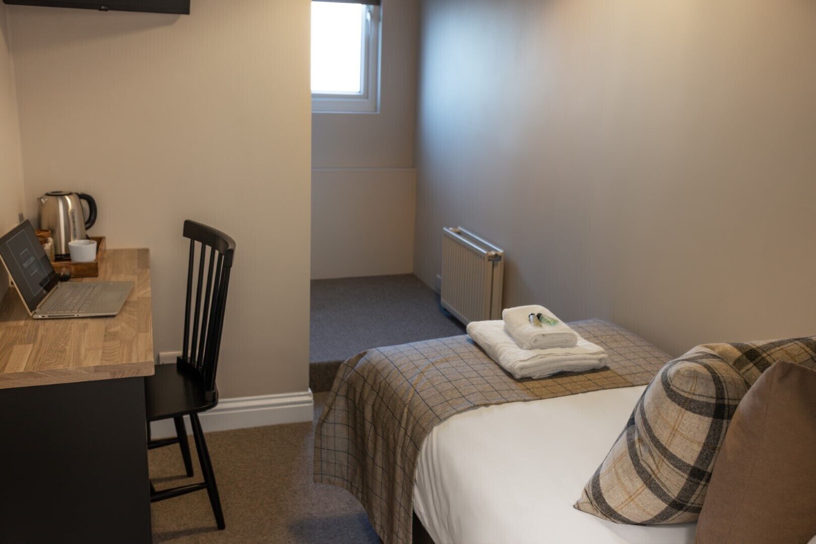 A small, neatly arranged room with a bed, a chair, a desk with a laptop, a kettle, and a radiator.