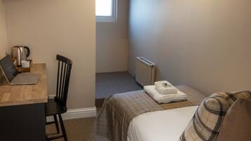 A small, neatly arranged room with a bed, a chair, a desk with a laptop, a kettle, and a radiator.