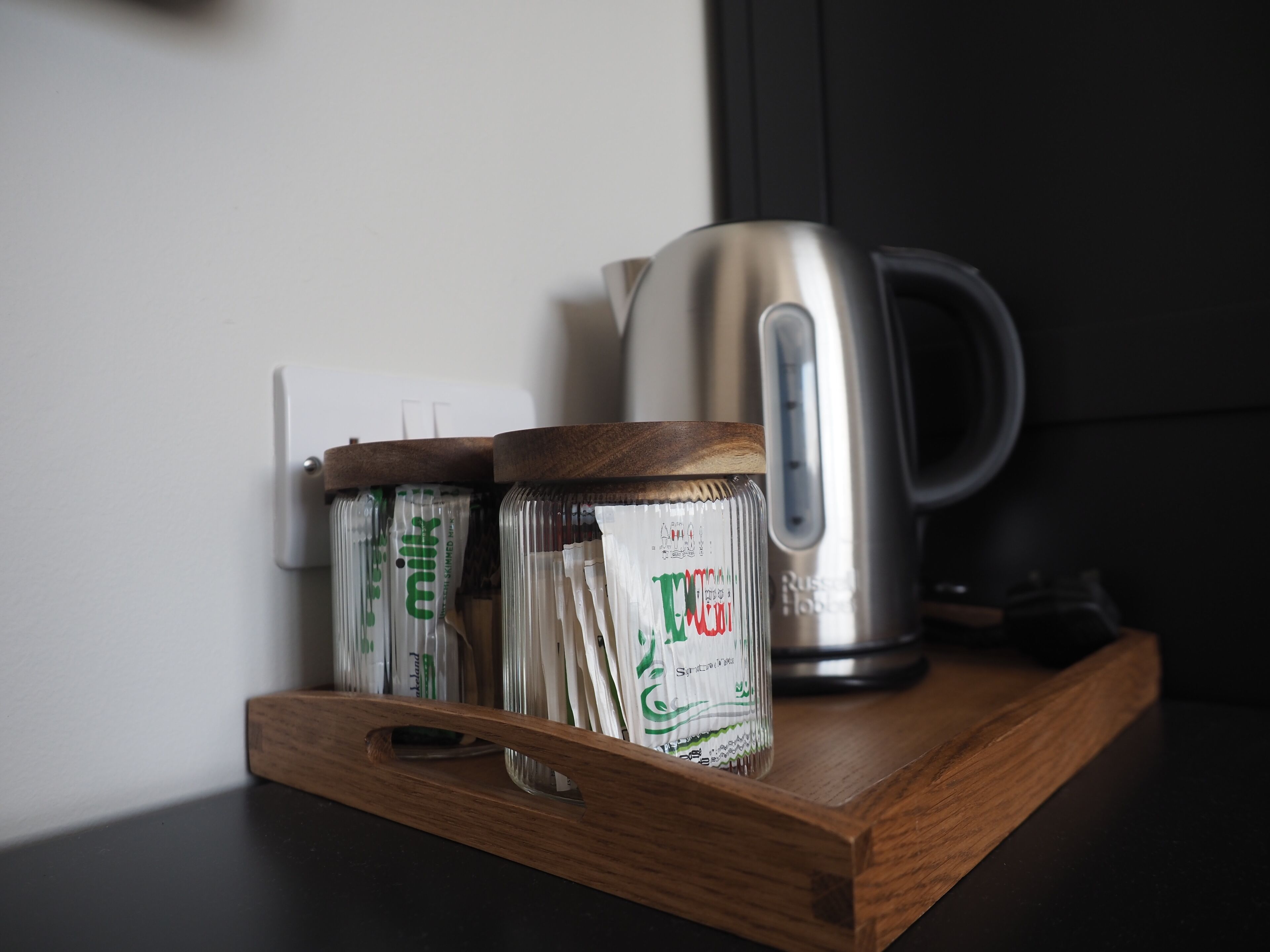 A wooden tray holding two jars with labels, a kettle, and a power outlet on a dark countertop.