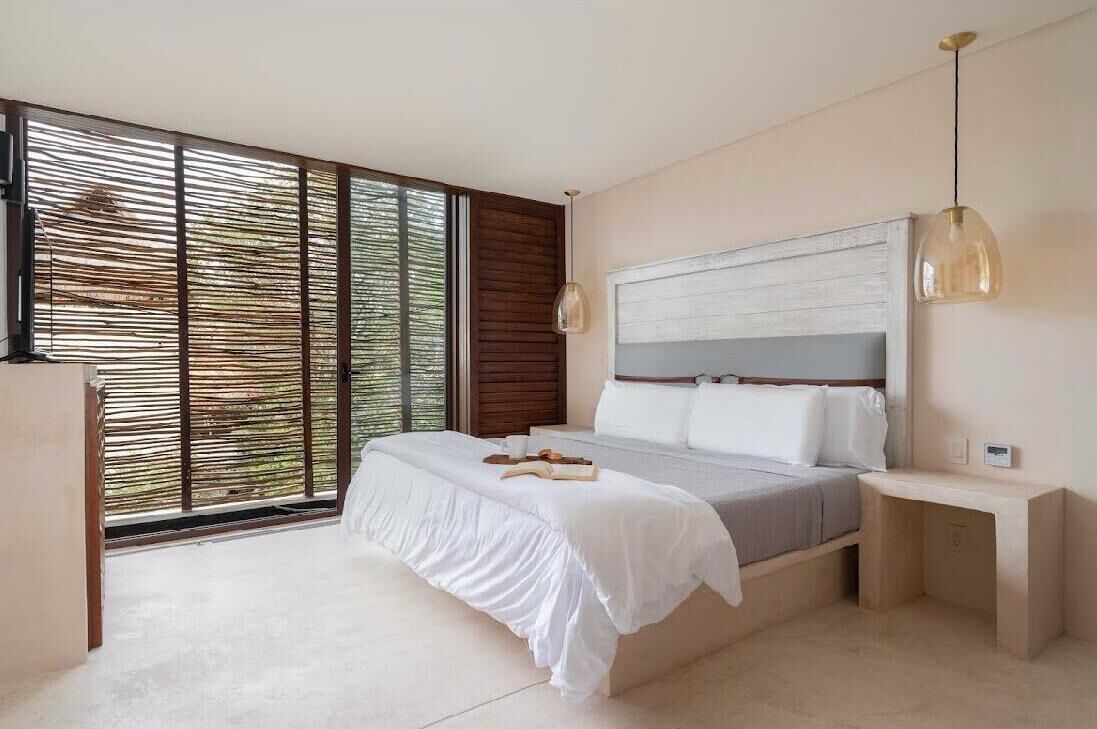 A modern bedroom with a large bed, bedside tables, a hanging light fixture, and a view through a window with blinds.