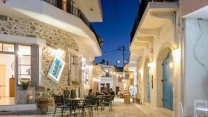 An outdoor café area with stone flooring and tables, surrounded by buildings with balconies and doors. - Leny's Seafront House (Panormos)