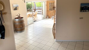 A room with a tiled floor, a wooden barrel, a wicker chair, and a view of an outdoor patio with a wooden cabinet and a glass door. - Villa Colombe by Agence Bru - 453196 (Carcans)