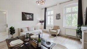 A modern living room with a sofa, armchair, dining table with wine glasses and a bottle, and a large window with curtains. - Vintage Apartment Bauhaus| 2 Bed Rooms| Netflix (Dessau-Roßlau)