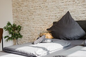 En pænt redt seng med gråt og hvidt sengetøj, en mørkegrå pude og et sammenrullet håndklæde. Et trænatbord med en lampe og en lille plante står ved siden af sengen.