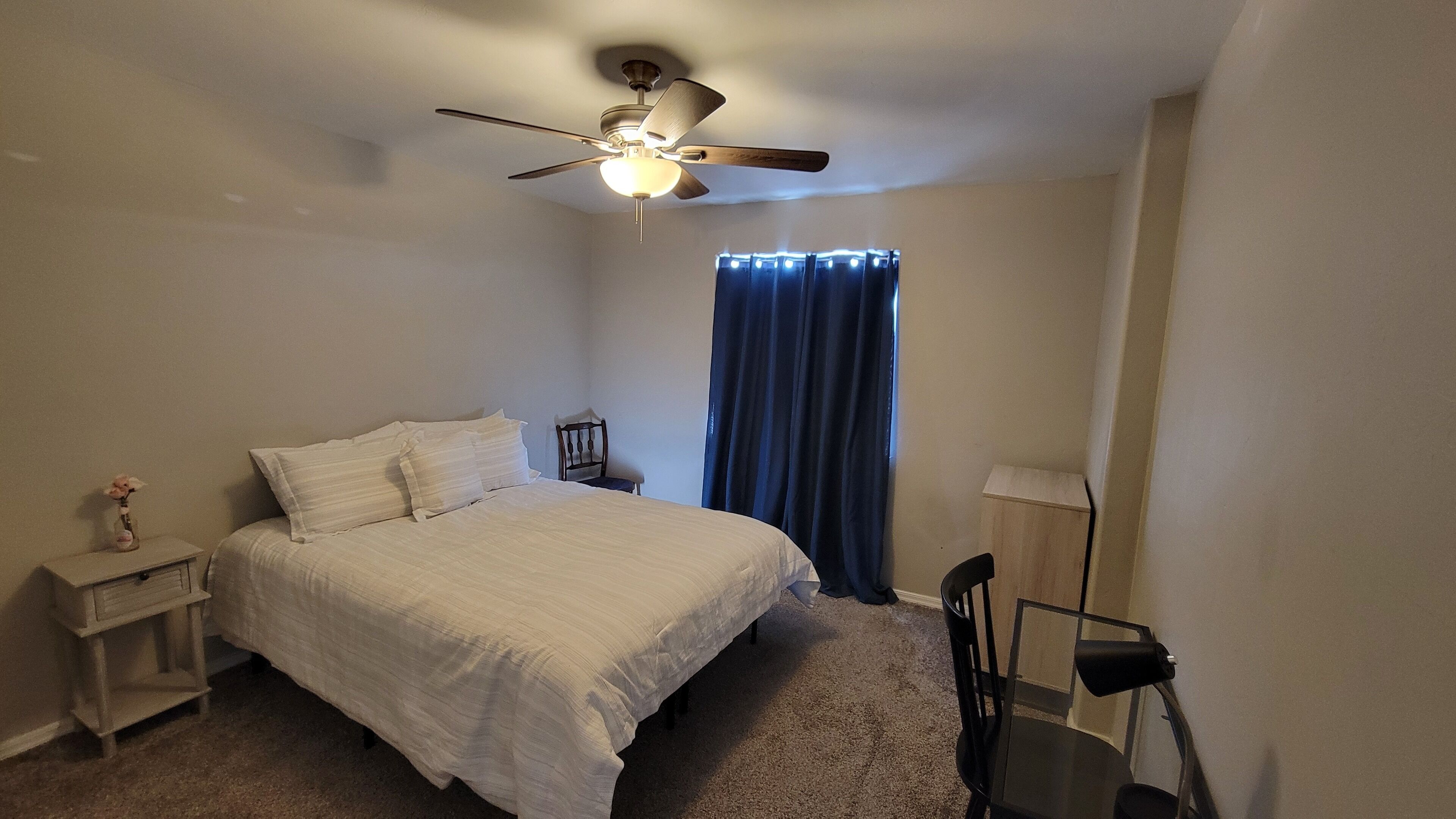 Une chambre à coucher comprenant un lit, une table de chevet, une chaise, un bureau, un ventilateur de plafond et une fenêtre avec des rideaux.