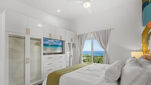 A bedroom with a bed, a TV, a wardrobe, and a view of the ocean. - Endless Island Horizons @ Coral Views, French Harbor (French Harbor)