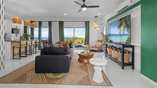 A modern living room with a sofa, coffee table, and TV, a kitchen area with bar stools, and a large window with a view. - Endless Island Horizons @ Coral Views, French Harbor (French Harbor)