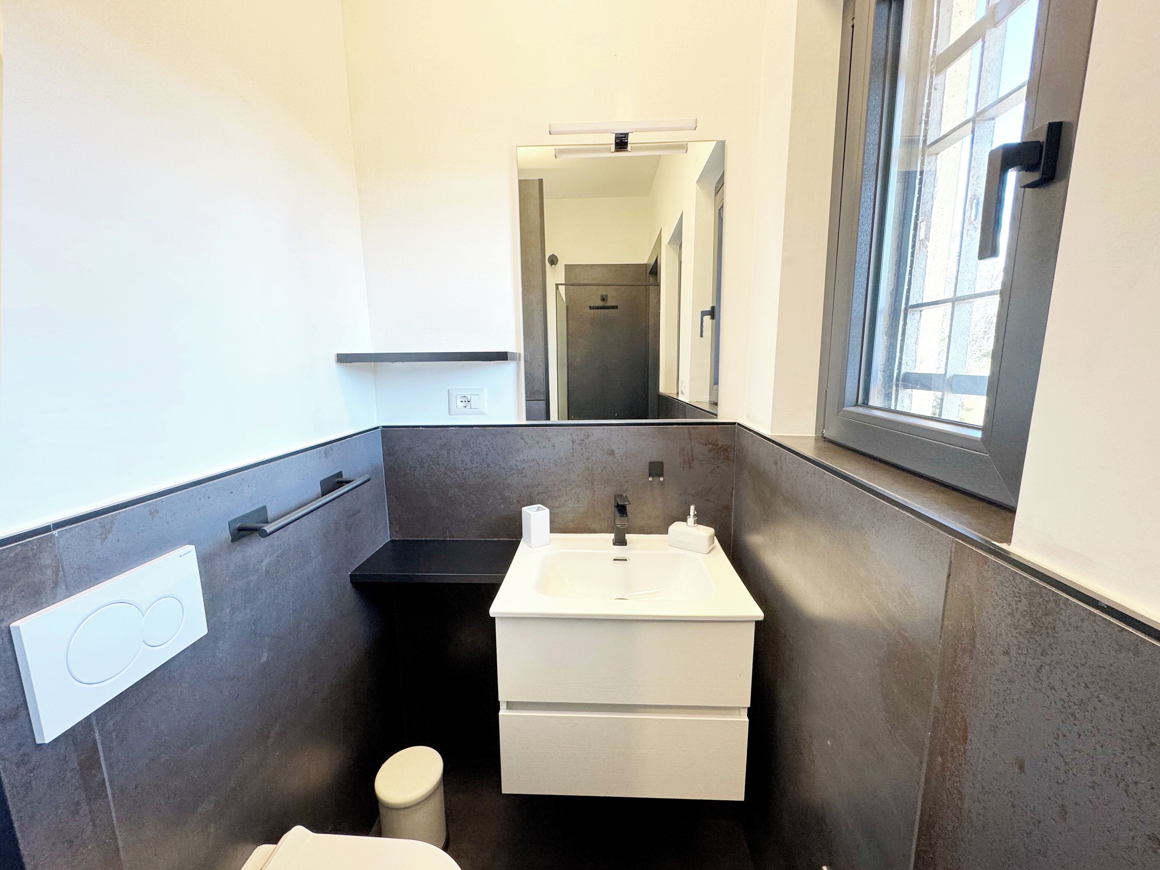 A modern bathroom with a white sink, dark tiled walls, a small toilet, and a window.