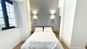 Ein ordentlich bezogenes Bett in einem Zimmer mit Fenster, zwei Wandleuchten und einem kleinen Nachttisch aus Holz.
