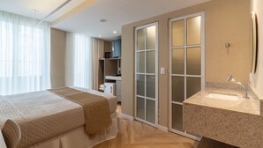 Une chambre d’hôtel moderne avec un grand lit, une salle de bain avec un lavabo et un miroir, et un placard avec une porte coulissante en verre.