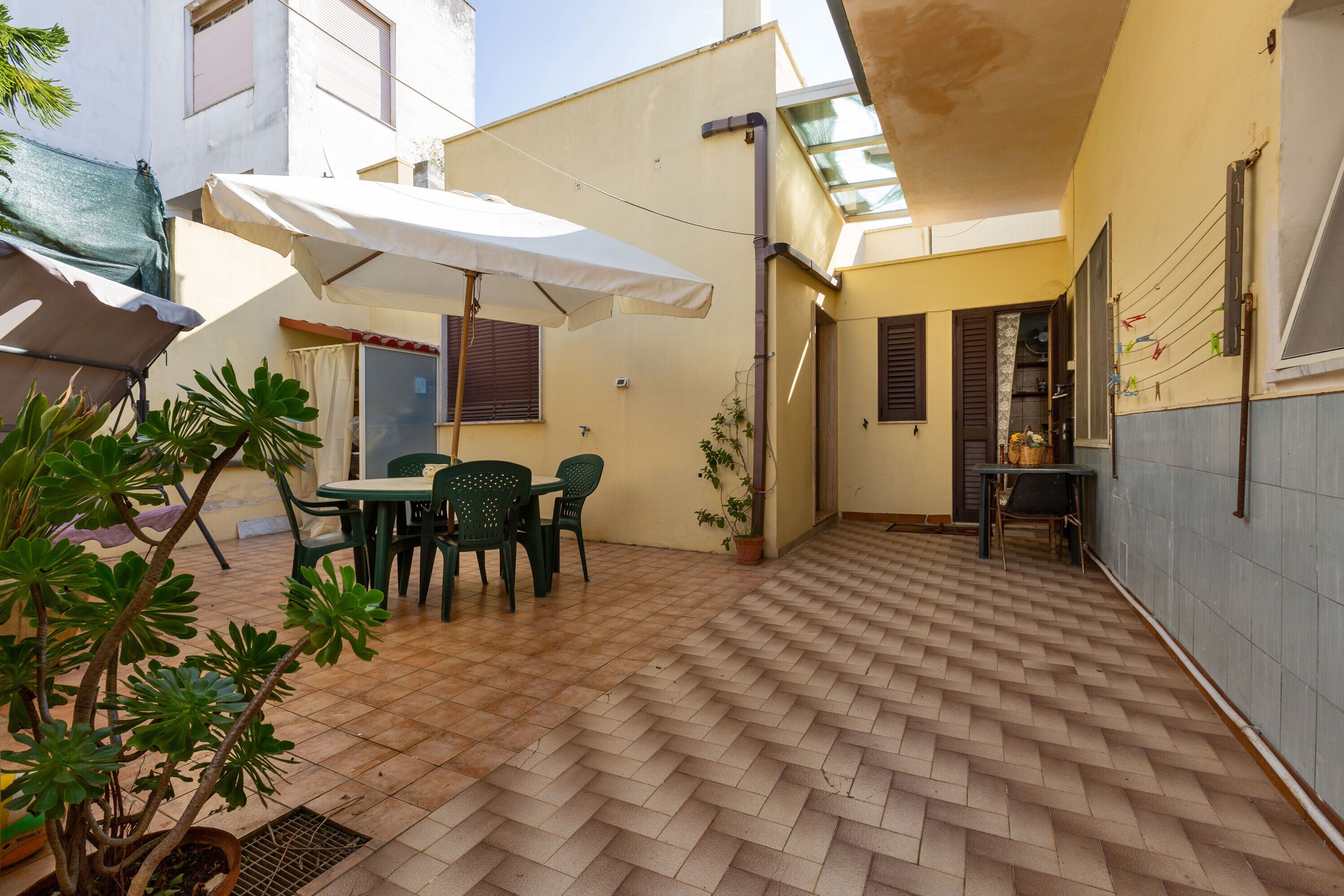 Une cour avec un sol carrelé, une table entourée de chaises, une plante en pot et un parasol blanc.