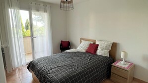 A bedroom with a bed, a nightstand, a lamp, and a view of the outdoors through a sliding door. - Apartment with large terrace, close to beaches, center and tourist sites! (Hyères)
