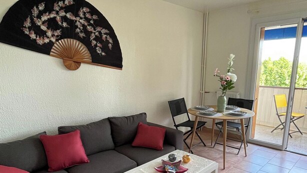 A living room with a grey sofa, a white coffee table, and a black fan with floral embroidery. There is a dining table with chairs and a vase of flowers. - Apartment with large terrace, close to beaches, center and tourist sites! (Hyères)