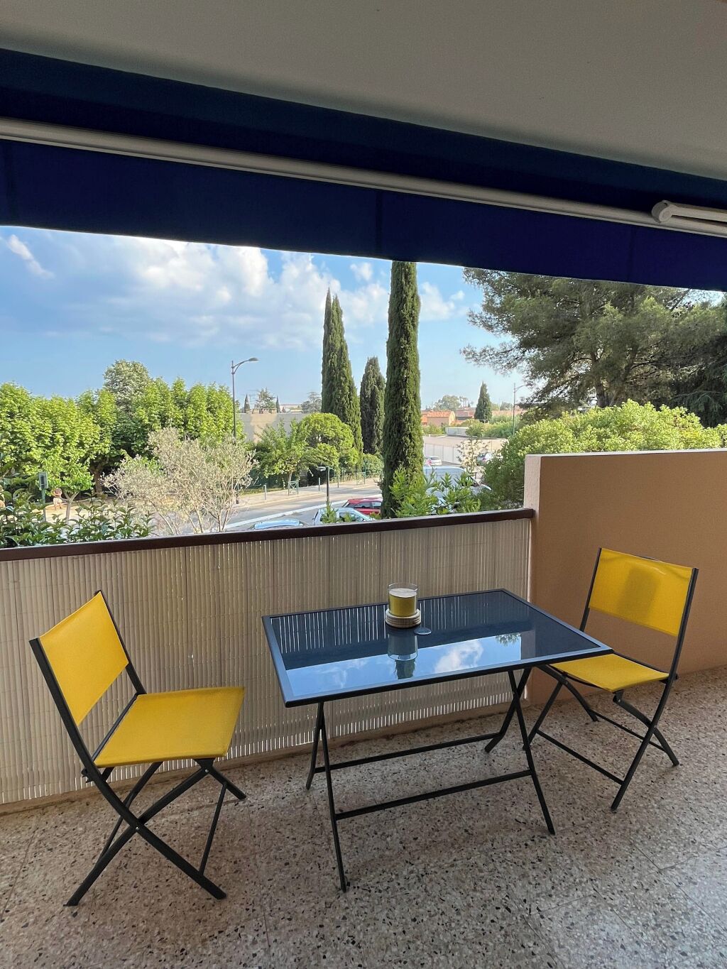 Un balcón con dos sillas amarillas y una mesa de vidrio, con vista a un estacionamiento y vegetación.