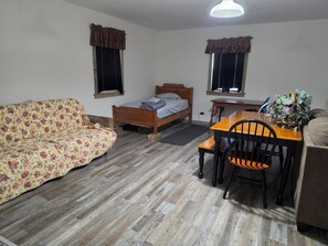 A room with a bed, a sofa, a dining table with chairs, and two windows with curtains.