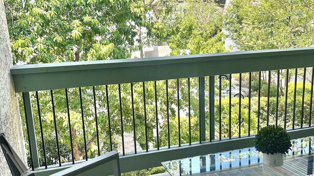 Ein Balkon mit grüner Geländer, einem Glastisch, einer gewebten Matte und einem Blumentopf.