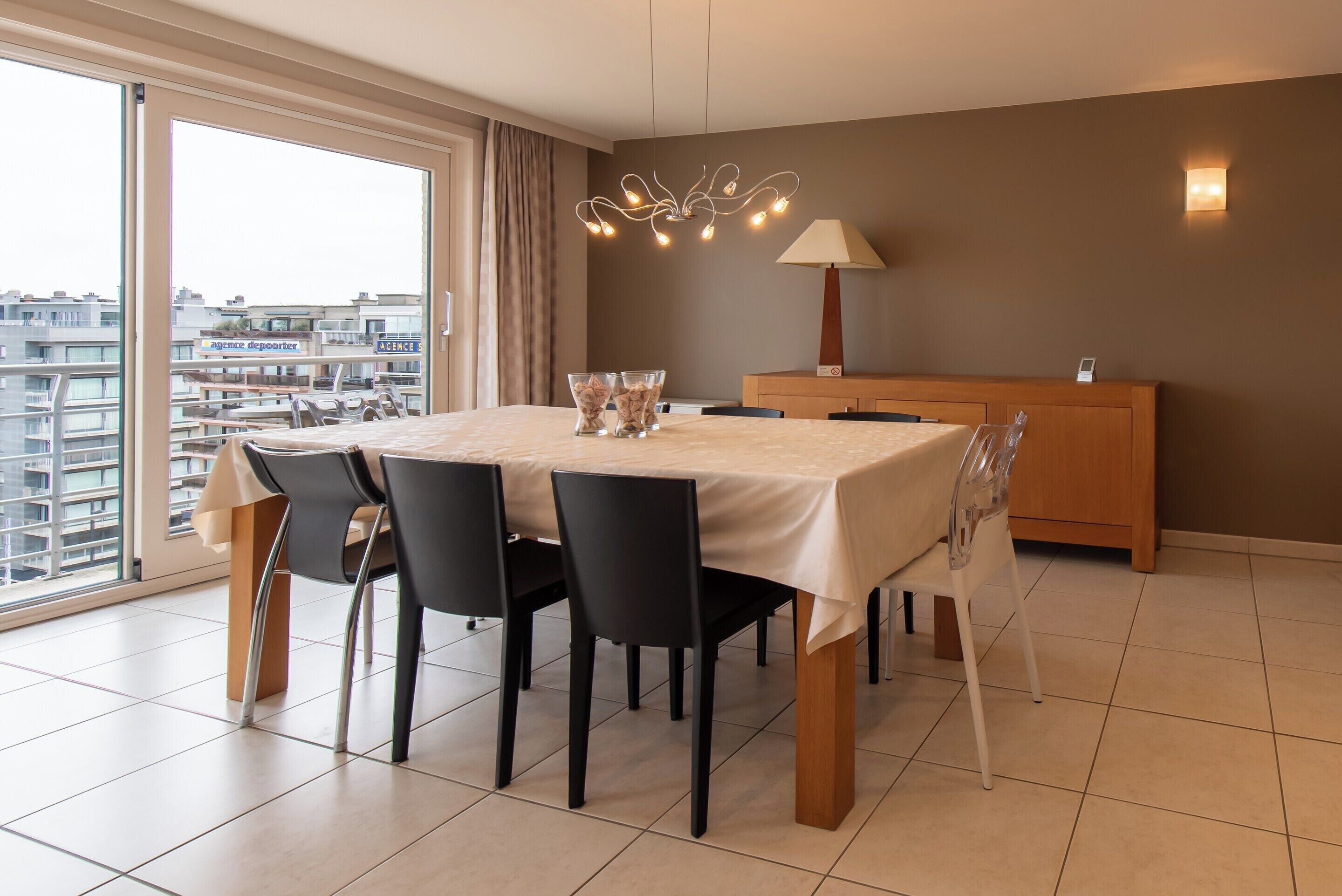 Uma sala de jantar moderna com uma mesa posta para quatro pessoas, um aparador de madeira e vista para a cidade através de grandes janelas.