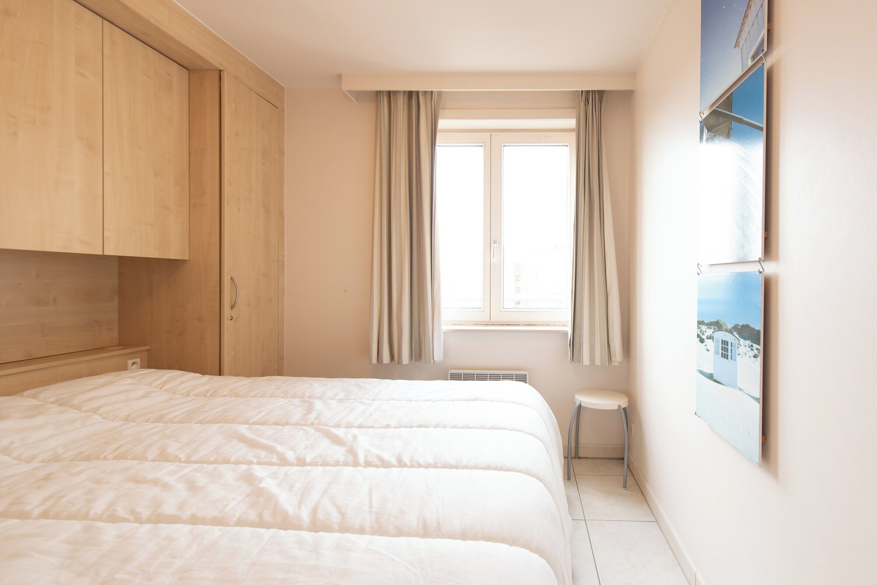 A bedroom with two beds, wooden wardrobe, a small table, and a window with curtains.