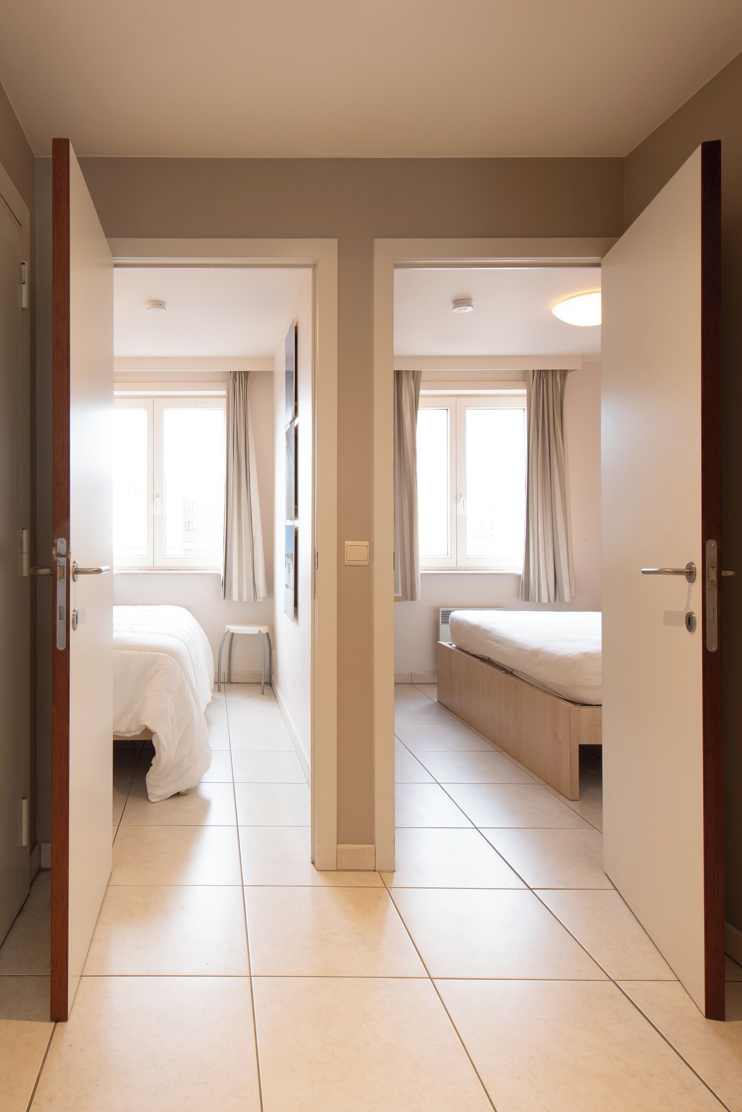 Ein Hotelzimmer mit Bett, einem kleinen Hocker und einem Fenster mit Vorhängen.