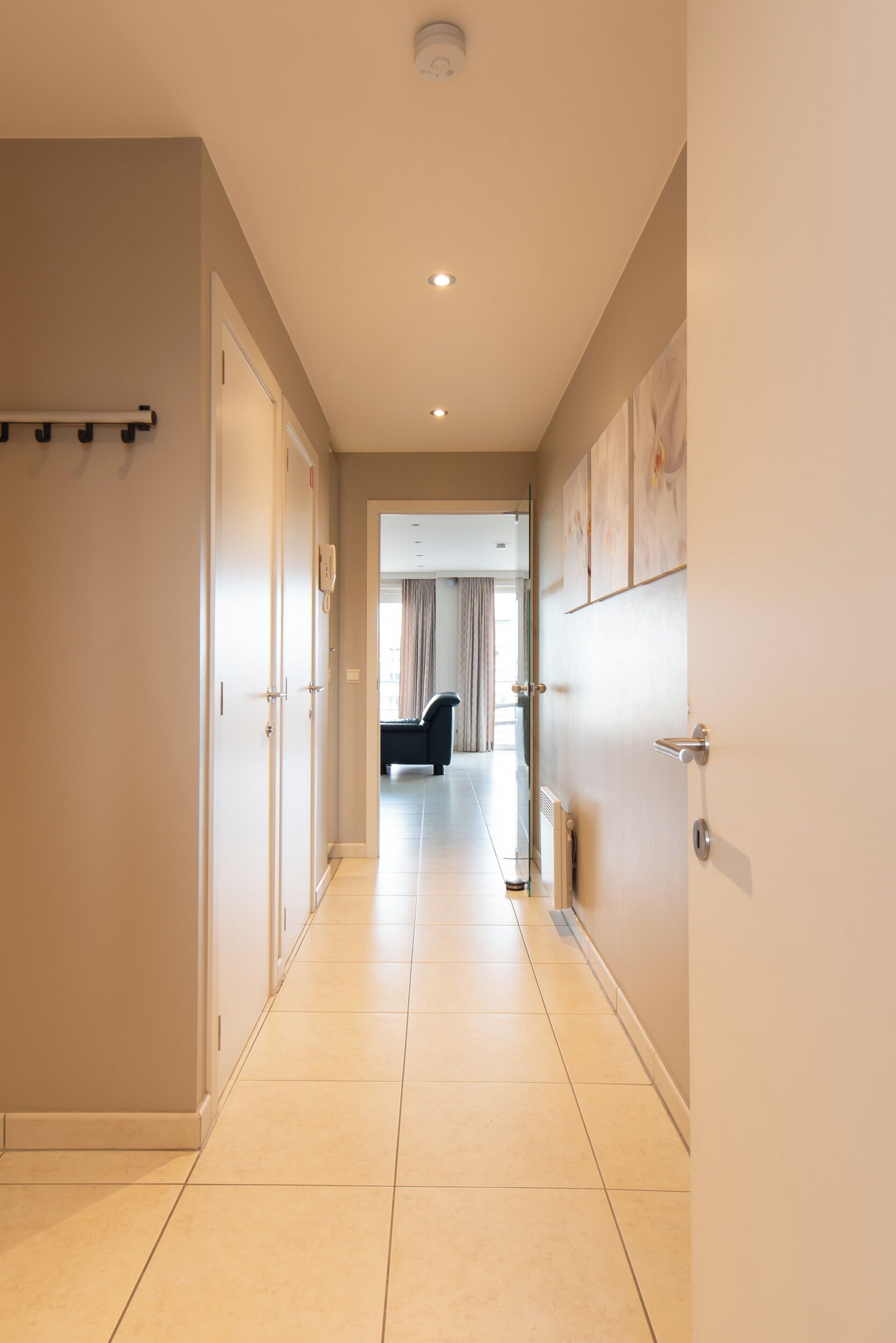 A hotel hallway with a long, tiled floor, a black sofa in the distance, and a door with a towel rack on the left.