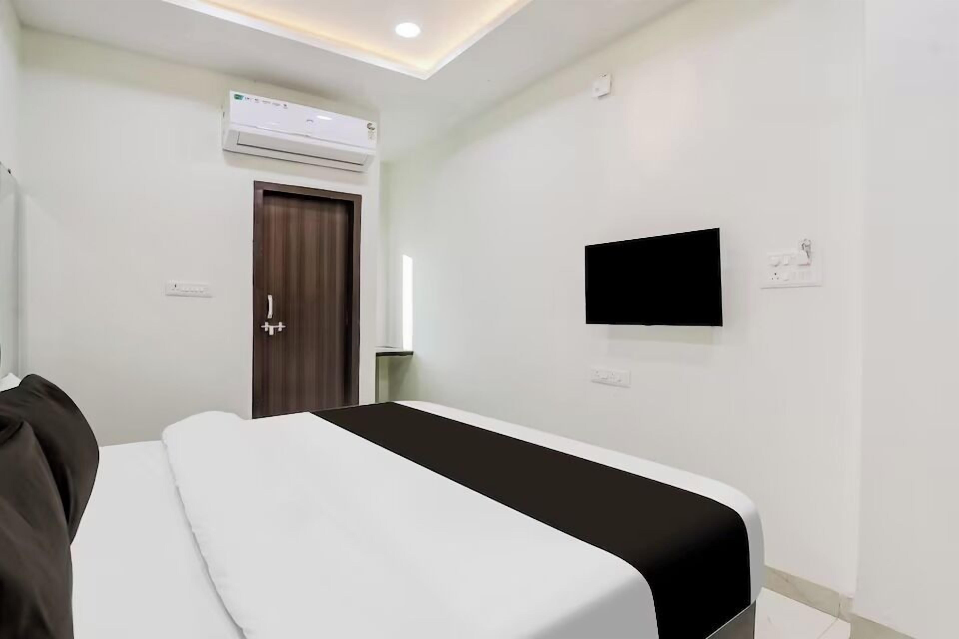 A hotel room with a bed, a flat-screen TV mounted on the wall, an air conditioning unit above the door, and a window with curtains.