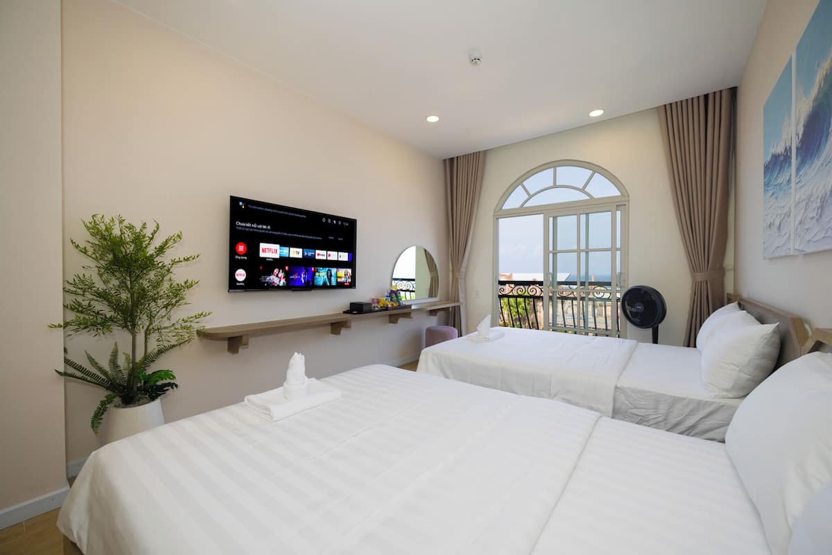A hotel room with two beds, a TV mounted on the wall, a potted plant, a window with a view, and a balcony with a railing.
